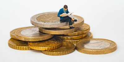 A stack of gold and silver coloured Euro coins with a small person wearing a blue jacket perched on the edge, reading a newspaper