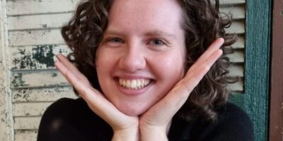 Photograph of a young white woman with curly brown hair. She is smiling as she props her face in her hands.